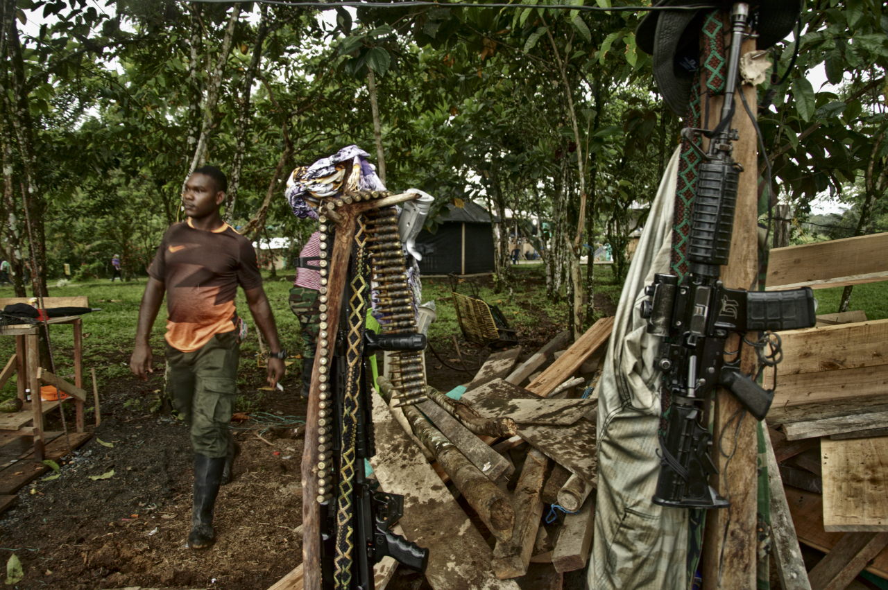 Una ametralladora con su munición y un fusil colgado de una estaca, al fondo dos hombres desarmados. Foto: Andrés Gómez.