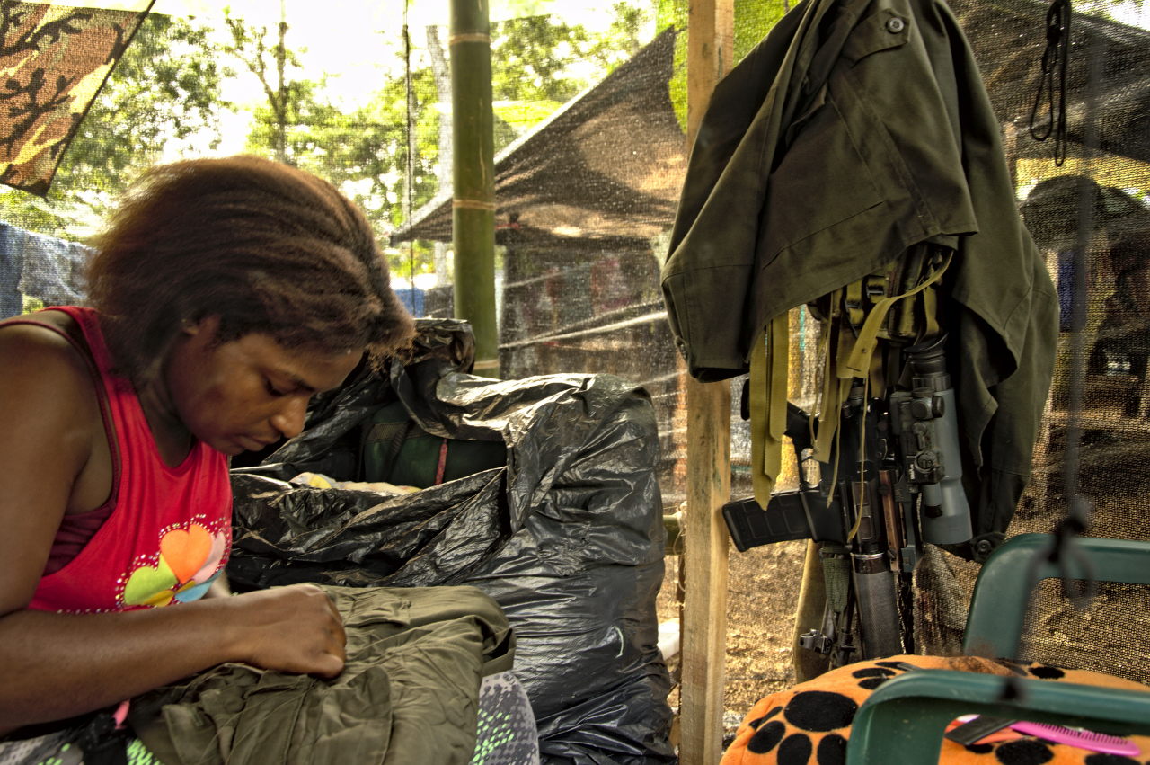 Una mujer cose un pantalón mientras su fusil está colgado de una estaca. Foto: Andrés Gómez.