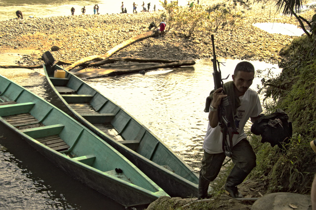 Un excombatiente sale de una lancha con su fusil en la mano, al fondo otros excombatientes se bañan en el río. Foto: Andrés Gómez.