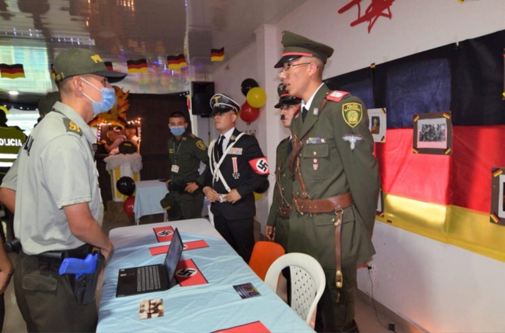Cinco policías, tres de ellos disfrazados como nazis. Al centro, una mesa con banderas de papel del Partido Nacional Socialista alemán; a la derecha, una bandera alemana.  