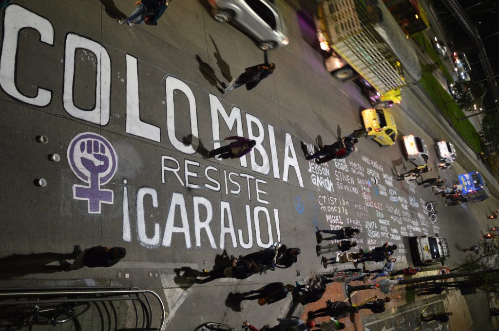 Una avenida, de un lado vehículos y de otro manifestantes. Entre ellos un letrero en el piso que dice: "Colombia resiste, ¡carajo!", seguido de un listado de nombres. Foto: Andrés Gómez