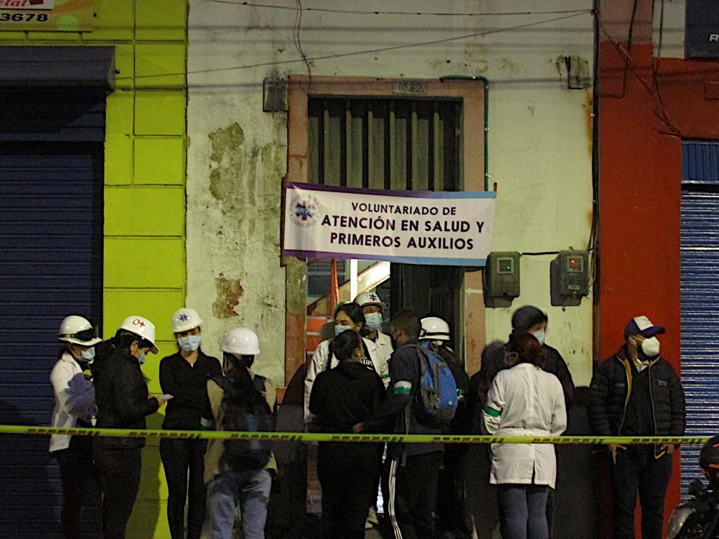 Un grupo de personas con cascos o batas blancas se reúne frente a la entrada de un edificio. Arriba de la puerta hay un cartel que dice: "Voluntariado de atención en salud y primeros auxilios". Foto: Andrés Gómez