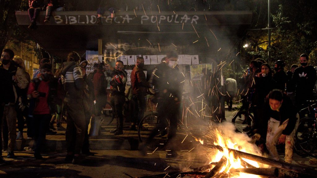 Un grupo de jóvenes se reúne del lado izquierdo de un fuego que una joven mantiene encendido. Al fondo una edificación de la Policía (CAI) con un grafiti que dice "biblioteca popular". Foto: Andrés Gómez