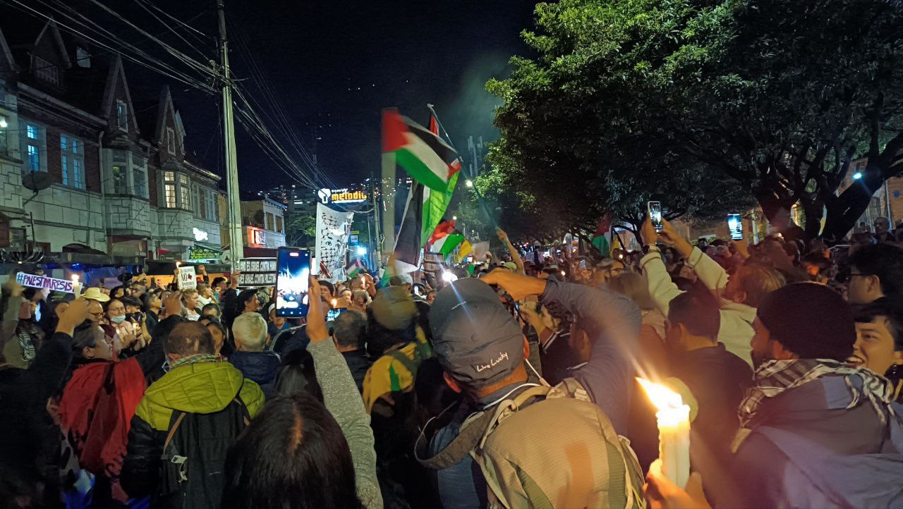 Una multitud se reúne en la noche junto a la embajada de Palestina en Bogotá, arriba se ven algunas banderas de ese país y en la esquina inferior derecha se ve a alguien portando unas velas encendidas. Foto: Joanna Zapata